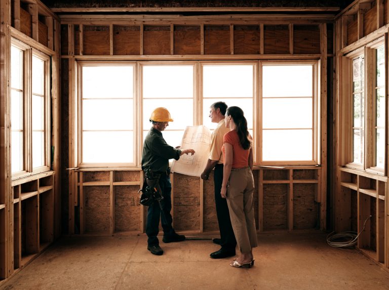 A man and woman look at blueprint plans with a contractor inside a room that's being remodeled.